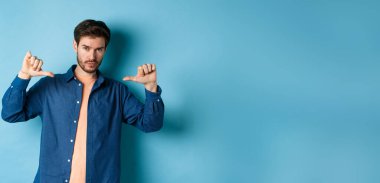 Sakallı yakışıklı ve kendinden emin bir adam, kameraya bakıyor ve kendini gösteriyor, kendini tanıtıyor, mavi arka planda duruyor..