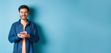 Kutlama ve tatil konsepti. Gülen adam elinde mumla doğum günü pastası tutuyor ve mutlu görünüyor, mavi arka planda duruyor..