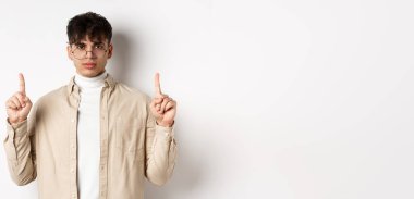 Gözlüklü yakışıklı hippi boş alanı işaret ediyor, kameraya ciddi bakıyor, beyaz arka planda duruyor..