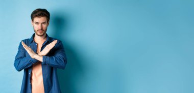 Ciddi beyaz adam hayır diyor, çapraz duruş hareketi yapıyor ve kameraya kendinden emin bakıyor, kötü bir şeyi yasaklıyor, mavi arka planda duruyor..