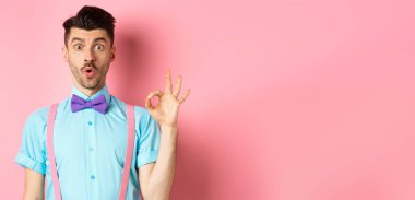 Papyonlu ve pantolon askısı takmış etkileyici bir adam hayret verici bir jest yapıyor, harika bir şey, pembe arka planda duruyor..