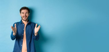 Neşeli genç adam kameraya el kaldırıyor ve gülümsüyor, senin için seviniyor, iyi iş çıkarıyor, mavi arka planda duruyor..