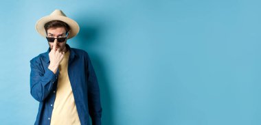 Yakışıklı ve arsız adam güneş gözlüğü ve hasır şapka taktı, yaz tatiline ya da tropikal tatil için hazır bekliyordu, mavi arka plan..