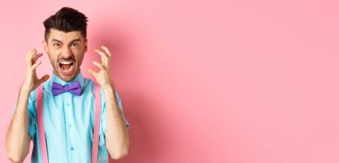 Frustrated young man losing temper, screaming and looking angry at camera, shaking hands pissed-off, standing mad on pink background.