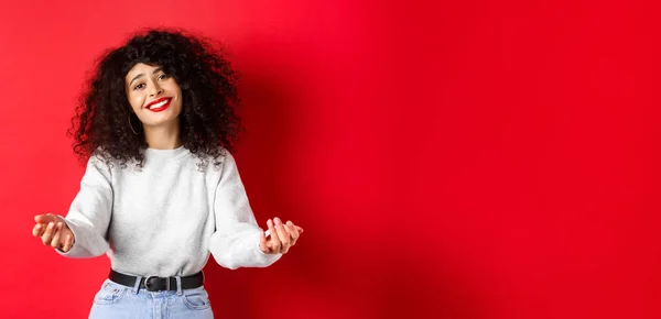 Pretty woman with curly haircut, extending hands and smiling with love and tenderness, inviting to come closer, reaching arms for hug, taking something into her arms, red background.