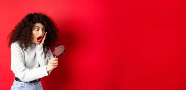Shocked girl look through magnifying glass with dropped jaw, seeing something amazing, standing on red background.