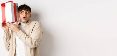 Tatil konsepti. Gözlüklü komik genç adam hediye kutusunu sallıyor, içerideki hediyeyi dinliyor ve hayrete düşürüyor, beyaz arka planda duruyor..