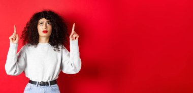 Endişeli, kıvırcık saçlı, kaşlarını çatmış ve şüpheli görünen, tereddütlü ya da endişeli yüzüyle kırmızı arka planda duran genç bir kadın..