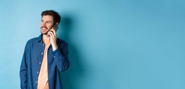 Yakışıklı beyaz adam cep telefonuyla konuşuyor, boş yere bakıyor kaygısız bir gülümsemeyle, mavi arka planda duruyor..