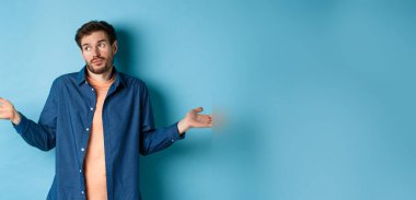 Sakallı kararsız genç adam, yüzünü çevirmiş, omuzlarını silkiyor, hiçbir şey bilmiyor, bilgisiz, mavi arka planda duruyor..