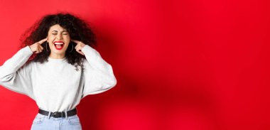 Sinirli kıvırcık kız parmaklarıyla kulaklarını tıkıyor ve bağırıyor. Kırmızı arka planda duran korkunç gürültüden rahatsız oluyor..