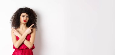 Kırmızı elbiseli, şüpheli ve üzgün genç bir kadın, parmaklarını yana çevirip somurtuyor, ürünler arasında karar veremiyor, seçimlerde yardıma ihtiyacı var, beyaz arka planda duruyor..