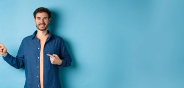 İlan veren mutlu beyaz adam, boş alanı işaret ediyor, mavi arka planda duruyor..