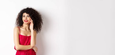 Kırmızı elbiseli ve makyajlı zarif ve romantik bir kadın. Gülümsüyor ve yüzüne dokunuyor. Mutlu bir ifadeyle, beyaz arka planda..