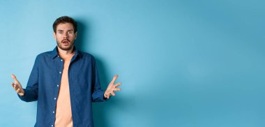 Şok olmuş genç adam ellerini kaldırıyor ve kameraya bakıyor. Korkmuş, inançsızlıkla bakıyor, mavi arka planda duruyor..