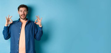 Etkileyici bir adam harika bir şeyi övüyor, iyi işaretler gösteriyor ve şaşırmış görünüyor, iltifat ediyor, mavi arka planda duruyor..