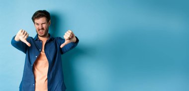 Tiksinç adam olumsuz görüş bildiriyor, baş parmaklarını aşağı ve dillerini gösteriyor, üzgün, kötü bir şeyi onaylamıyor, mavi arka planda duruyor..