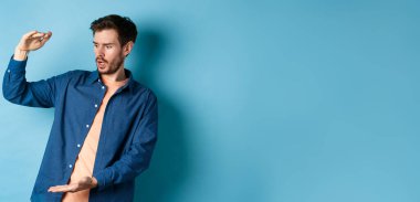 Şok olmuş genç adam boş alana bakıyor ve mavi arka planda duran büyük boyutlu nesneleri gösteriyor..