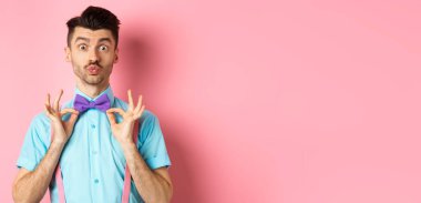 Yakışıklı ve komik genç adam öpüşmek için dudaklarını büzüyor, şık papyonunu gösteriyor, parti için kıyafetler giyiyor, pembe arka planda duruyor..