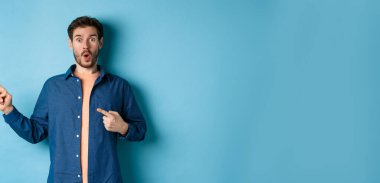 Şaşırmış ve şaşırmış beyaz adam hayranlıkla soluk soluğa kalmış, sol logoyu işaret ediyor, müthiş bir tanıtım yapıyor, mavi arka planda duruyor..