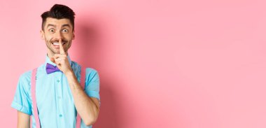 Neşeli genç adam sürpriz yapıyor, mutlu bir gülümsemeyle kameraya şşşş yapıyor, sesini alçaltmasını istiyor, sessiz olmasını istiyor, pembe arka planda duruyor..