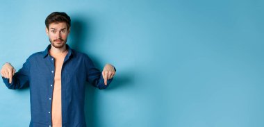 Şaşkın erkek manken gündelik giysiler içinde şaşkın şaşkın bakıyor ve işaret ediyor, afişi gösteriyor, mavi arka planda duruyor..