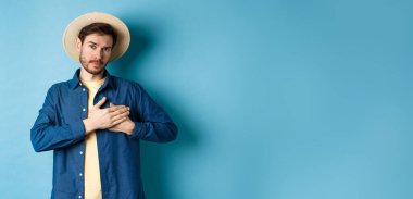 İçten bir genç kadın, turist şapkası takmış, elini kalbe koyuyor ve minnettar ve ciddi görünüyor, mavi arka planda duruyor..