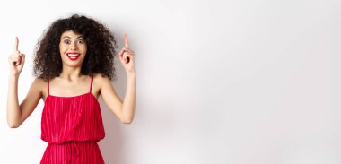 Kırmızı elbiseli ve makyajlı mutlu zarif bir kadın, gülümsüyor ve logoyu işaret ediyor, reklamını yapıyor, beyaz arka planda duruyor..