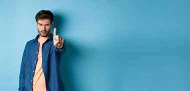 Ciddi genç adam seni durdurmak ya da uyarmak için bir parmağını uzatıyor, bir şeyi yasaklıyor ya da onaylamıyor, mavi arka planda duruyor. Boşluğu kopyala