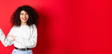 Kıvırcık saçlı, kırmızı dudaklı, gülümseyen beyaz kadın logoyu işaret ediyor, tanıtım teklifini gösteriyor, stüdyo geçmişine karşı duruyor..