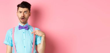 Papyon ve pantolon askısı takmış kafası karışmış, kendini suçlanmış ya da seçilmiş gibi gösteren, kaşlarını kaldıran, pembe arka planda duran komik adamın görüntüsü..