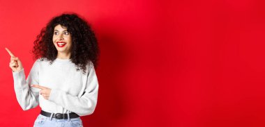 Heyecanlı, kırmızı dudaklı kıvırcık kadının portresi, logoyu şaşırmış bir şekilde gösteriyor, tanıtım anlaşmasına bakıyor, stüdyo arka planında duruyor..