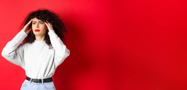 Sıkıntılı kıvırcık saçlı İtalyan kız, baş şakaklarına dokunuyor ve kötü hissediyor, baş ağrısı çekiyor, yana bakıyor, kırmızı arka planda duruyor..