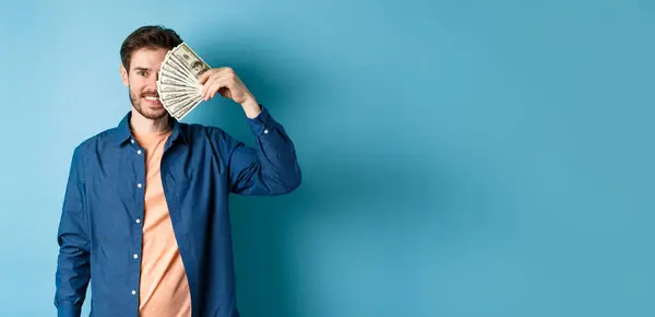 Happy young guy covering half of face with dollars and smiling, winning prize money, standing on blue background.