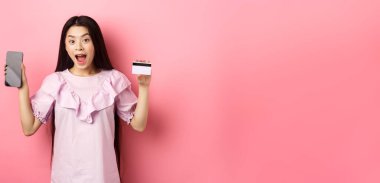 İnternetten alışveriş. Heyecanlı Asyalı kadın boş akıllı telefon ekranlı plastik kredi kartı gösteriyor. Reklam interneti, pembe arka planda duruyor..