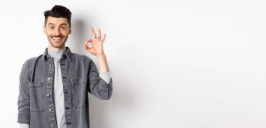 Gülümseyen yakışıklı adam iyi ürün gibi tamam işareti gösteriyor, reklam öneriyor, beyaz arka planda tatmin olmuş duruyor..