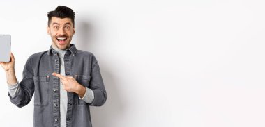 Heyecanlı yakışıklı adam boş akıllı telefon ekranı gösteriyor ve gülümsüyor, şuraya bakın, jest, beyaz arka planda duruyor..