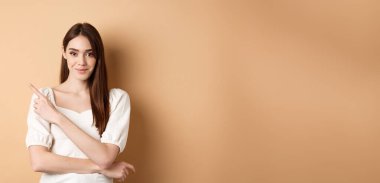 Beyaz elbiseli, gülümseyen güzel bir kız sol logoyu gösteriyor ve kendinden emin bir şekilde bej arka planda duruyor..