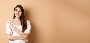 Beyaz elbiseli neşeli kız promosyon teklifini kontrol ediyor, sol üst köşeyi işaret ediyor ve rüya gibi bir gülümsemeyle, bej arka planda duruyor..