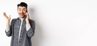 Heyecanlı adam cep telefonuyla konuşuyor ve iyi haberler duyuyor, seviniyor ve mutlu bir gülümsemeyle elini kaldırıyor, beyaz arka plana karşı duruyor..