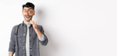 Mutlu komik adam büyüteçten büyük gözlerle bakıyor, ilginç bir anlaşmaya bakıyor, bir şeyler arıyor, beyaz arka planda duruyor..