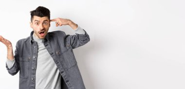 Delirdin mi sen? Sinirli bir adam aptalca davranışları için insanları azarlıyor, baş tapınağını işaret ediyor ve kafası karışmış bir şekilde elini kaldırıyor, beyaz arka planda dururken şikayet ediyor..