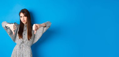 Olumsuz duygu. Uzun saçlı ve elbiseli, hayal kırıklığına uğramış bir kadın, baş parmaklarını aşağı gösteriyor ve suratını ekşitiyor, hoşnutsuzluğunu ifade ediyor, mavi arka planda duruyor..