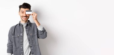 Bıyıklı mutlu yakışıklı adam gözlerinde plastik kredi kartı, kameraya heyecanlı bir şekilde gülümsüyor, beyaz arka planda duruyor..