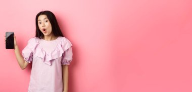 İnternetten alışveriş. Heyecanlı Asyalı kadın boş bir akıllı telefon ekranı gösterip pembe arka plana karşı durarak kameraya hayret içinde bakıyor..