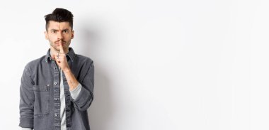 - Sessiz ol. Kızgın adam somurtuyor ve susuyor, sakin olmasını söylüyor, kötü davranışları azarlamak için parmaklarını dudaklarına bastırıyor, beyaz arka planda duruyor..