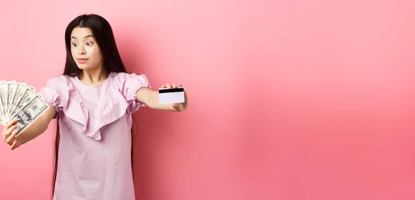 Excited teen asian girl stretch out hands with dollar bills and plastic credit card, look at money with amazement, standing on pink background.