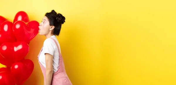 Valentines day and relationship concept. Profile of young asian woman scream of surprise, say wow and looking left amazed, standing near red balloons, yellow background.