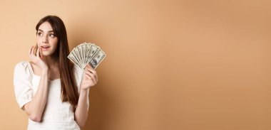 Hayalperest kız elinde dolarlarla alışverişe çıkıyor, sol üst köşeye bakıyor, düşünceli, parayı nasıl harcayacağını planlıyor, bej arka planda duruyor..