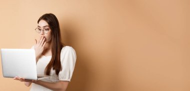Şok olmuş kız dizüstü bilgisayara bak, ağzı şaşkın bir ifadeyle kapla, bej arka planda gözlüklerle dur..